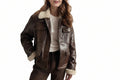 A woman wearing a dark brown leather jacket with a cream sherpa lining on the collar, cuffs, and pockets, paired with a cream ribbed sweater and dark brown pants, against a solid white background.